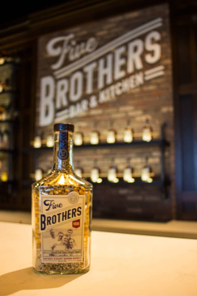 A bottle of Five Brothers brand bourbon sits, illuminated in the foreground with a large display of the bourbon in the background. 