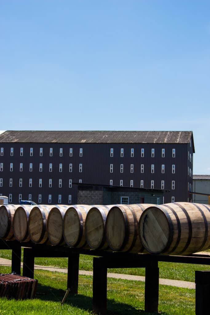 Barrels of bourbon sit on an outdoor display with a large rustic building in the background. The sky is blue and clear.