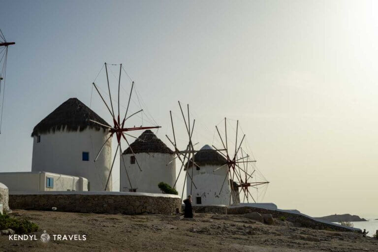 Historic windmills of Mykonos at sunset with a traveler standing nearby, overlooking the Aegean Sea in Greece.