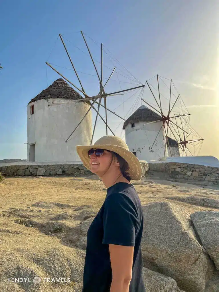 Smiling woman in a straw hat and sunglasses standing in front of the iconic windmills of Mykonos during golden hour.