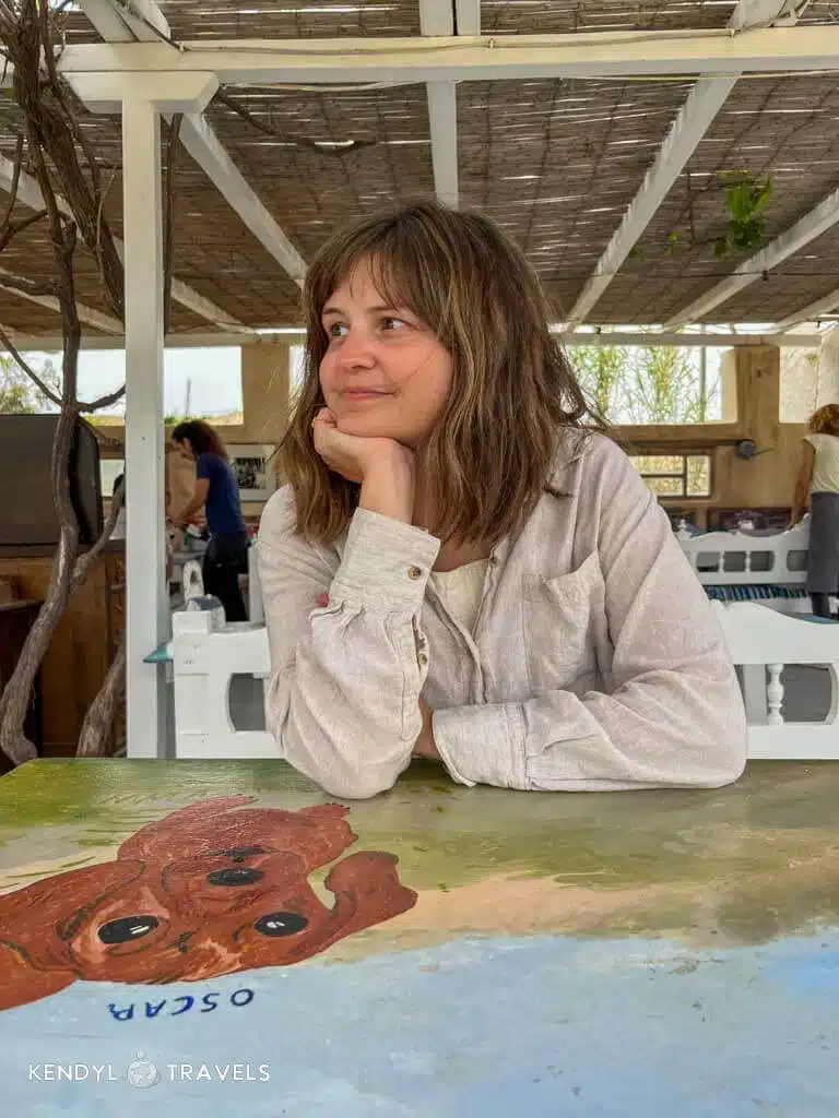 Woman sitting at a painted outdoor table at a rustic taverna in Greece, smiling and looking to the side under a shaded pergola.
