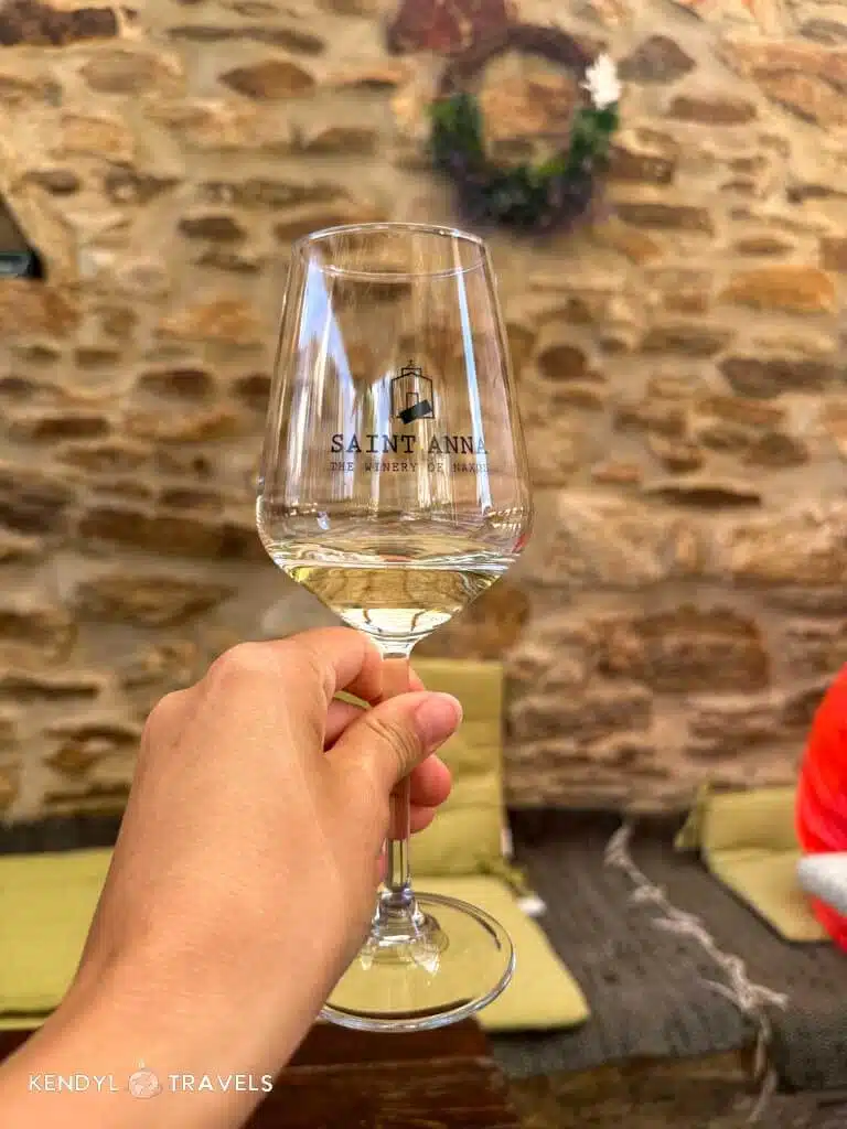 A hand holding a glass of white wine at Saint Anna winery in Naxos, with a rustic stone wall and cozy seating in the background.