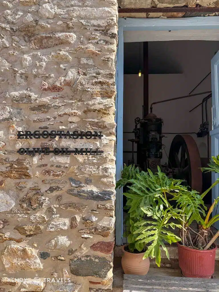 The stone exterior and entrance of the historic Ergostasion Olive Oil Mill in Naxos, with green potted plants flanking the doorway.