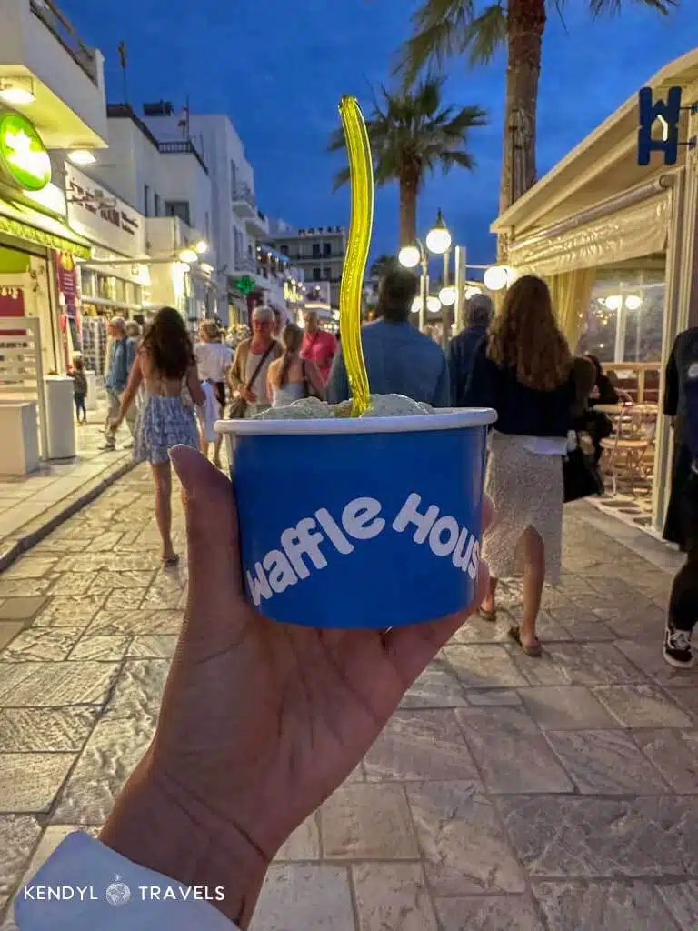 A hand holding a cup of gelato from Waffle House in Naxos, with a bustling, lantern-lit street scene in the background.