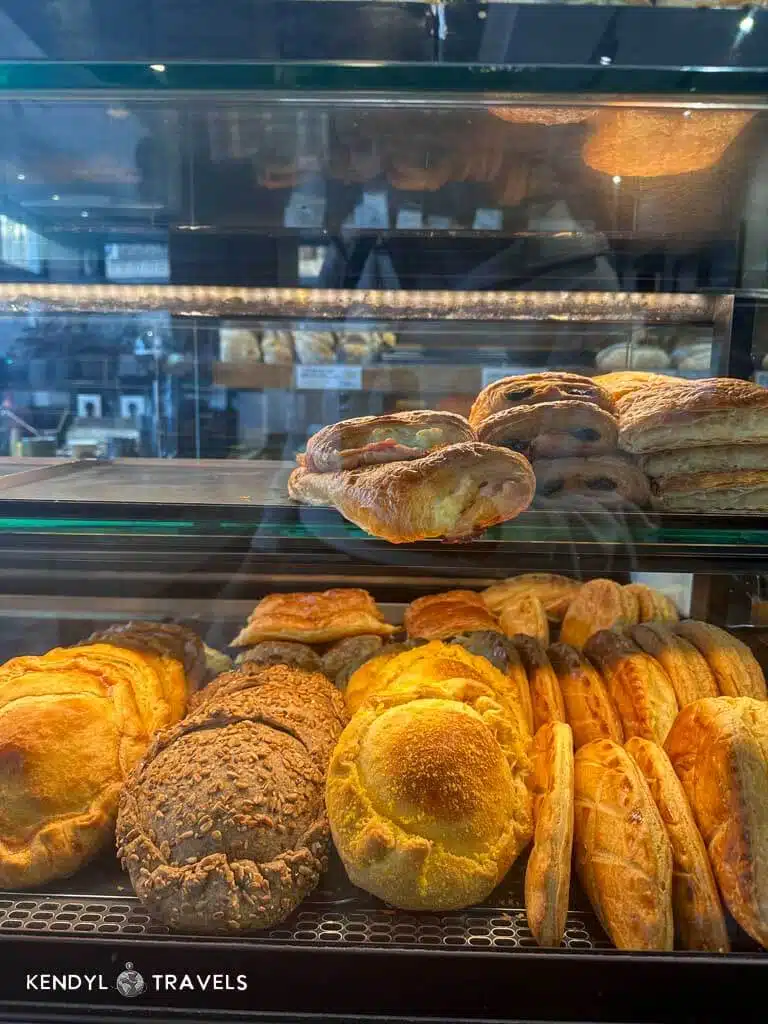 A bakery display case filled with fresh Greek pastries and breads, including seeded rolls, puff pastries, and golden-brown buns.