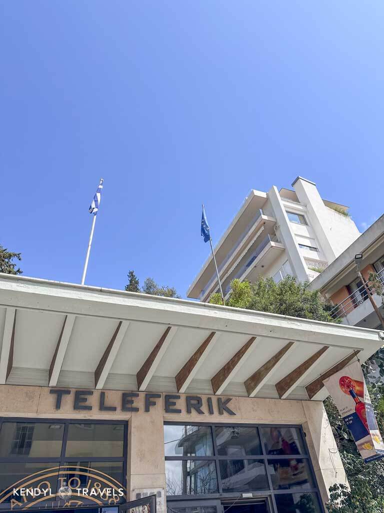 Entrance to the Teleferik (funicular) station in Athens on a clear blue day, with Greek and European Union flags flying above and a modern apartment building in the background.