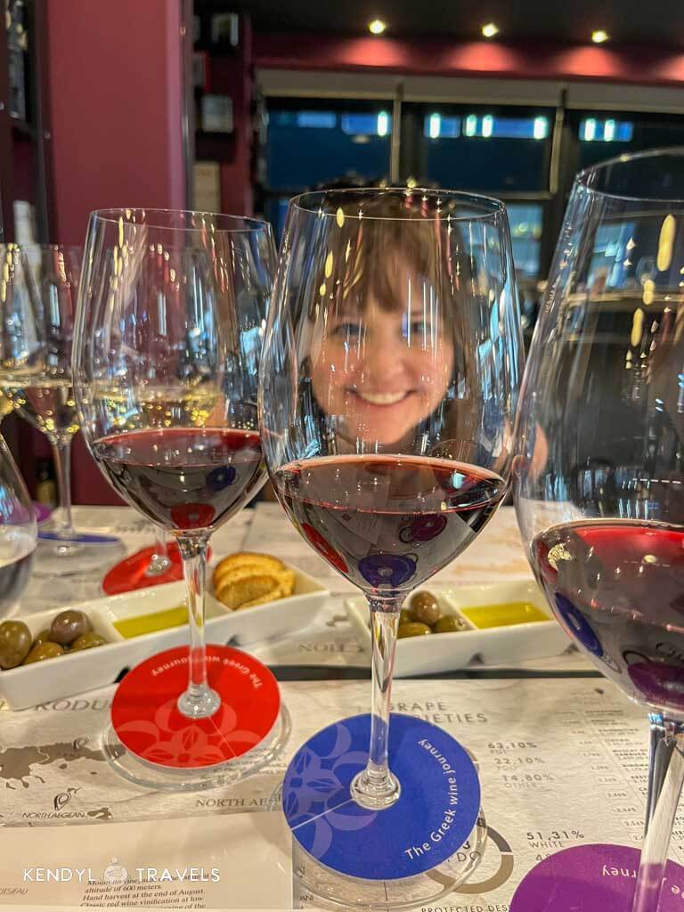 A smiling woman seen through a row of red wine glasses during a Greek wine tasting, with olives, bread, and tasting notes laid out on the table in a cozy, dimly lit setting.