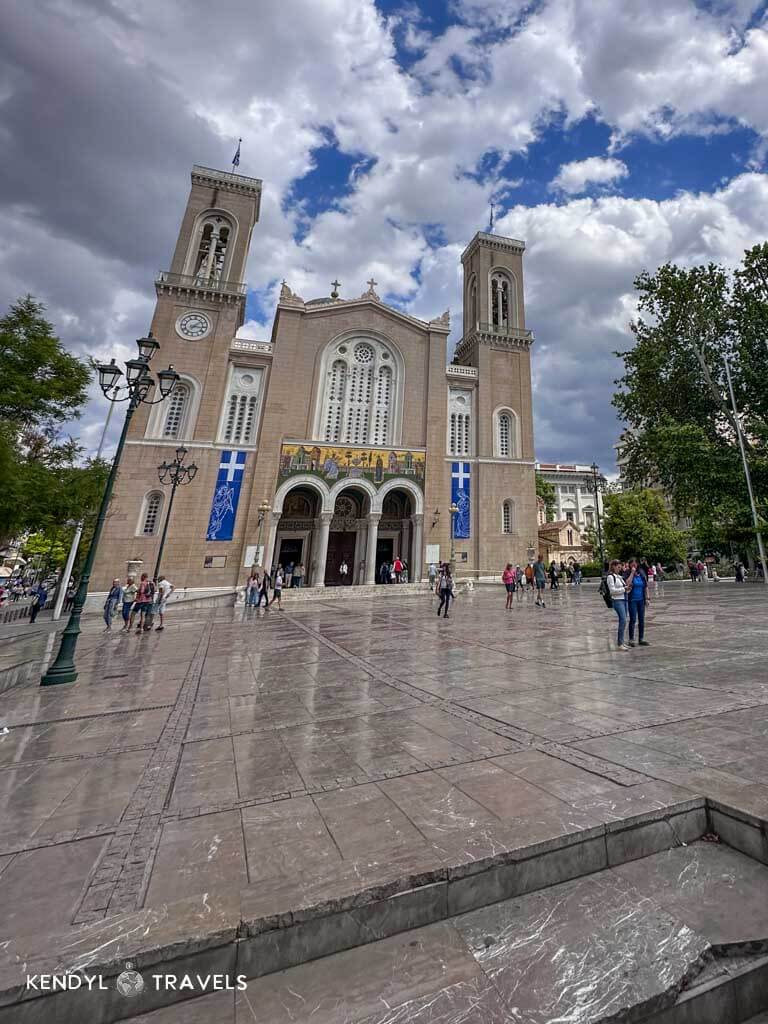 The exterior of the Metropolitan Cathedral of Athens, featuring twin bell towers and a wide marble plaza in front. The sky above is partly cloudy, and people are walking or gathering near the entrance beneath arched doorways and vibrant mosaics.