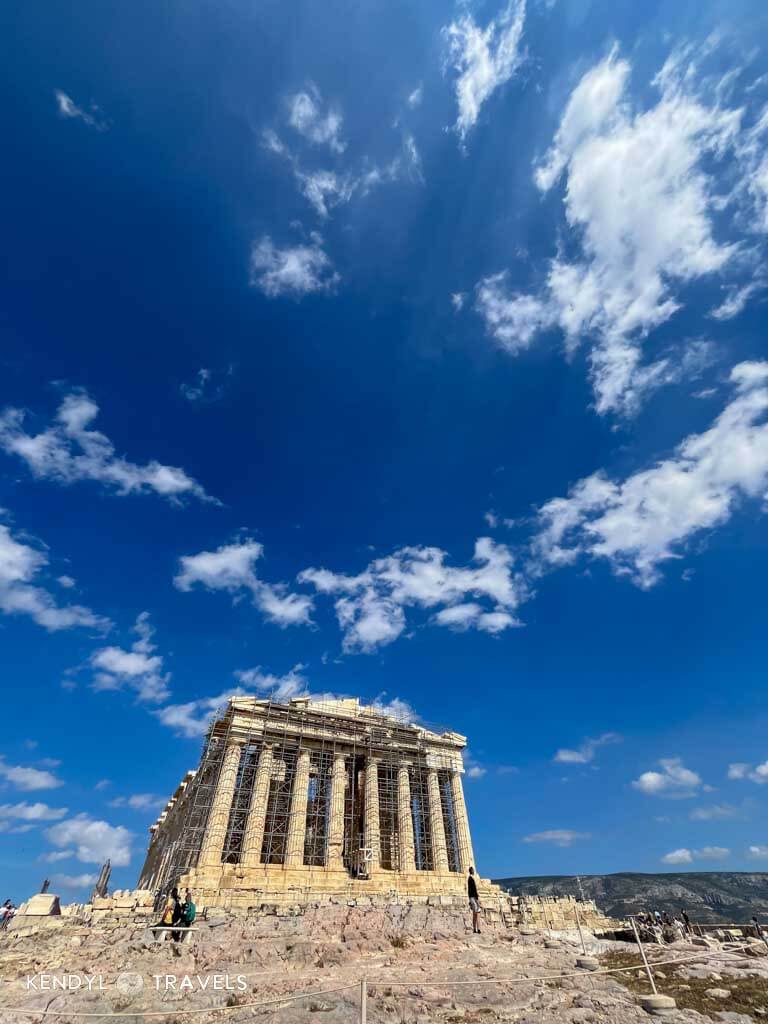 The Parthenon stands atop the Acropolis in Athens, surrounded by scaffolding for restoration under a vivid blue sky with scattered white clouds.