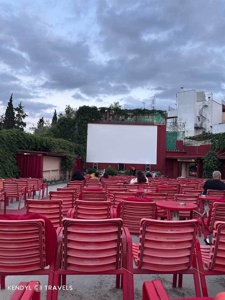 Rows of red chairs face a blank white movie screen at Thision Open Air Cinema in Athens, with a few people seated and the sky above filled with evening clouds.
