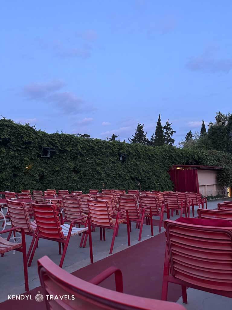 Empty red chairs line the outdoor seating area of Thision Open Air Cinema at dusk, surrounded by ivy-covered walls and silhouetted trees under a fading blue sky.