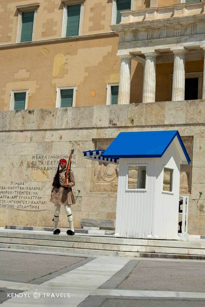 An Evzone guard in traditional uniform stands at attention beside a small white-and-blue sentry box outside the Hellenic Parliament in Athens, with stone carvings and shuttered windows in the background.
