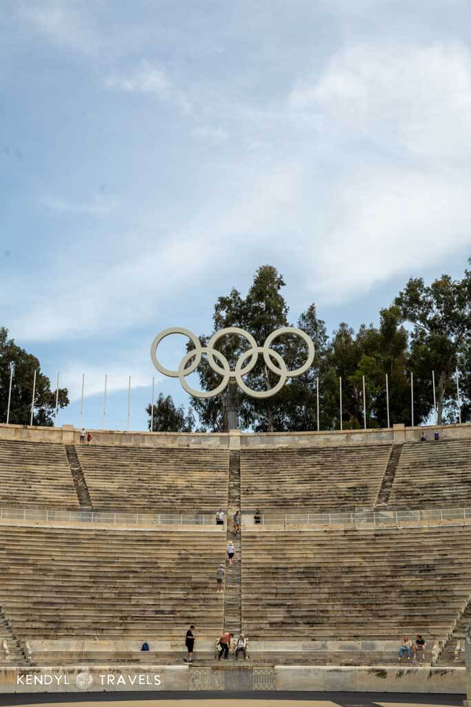 The marble seating of the Panathenaic Stadium in Athens, crowned with the Olympic rings and surrounded by tall trees under a lightly clouded sky, with visitors scattered across the central staircase.