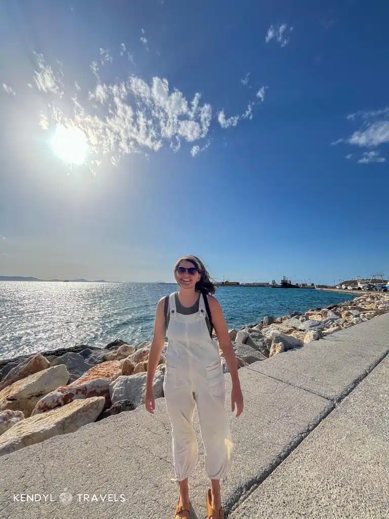 Woman wearing breezy overalls and sandals on a sunny seaside walk in Greece, an ideal outfit for May weather.
