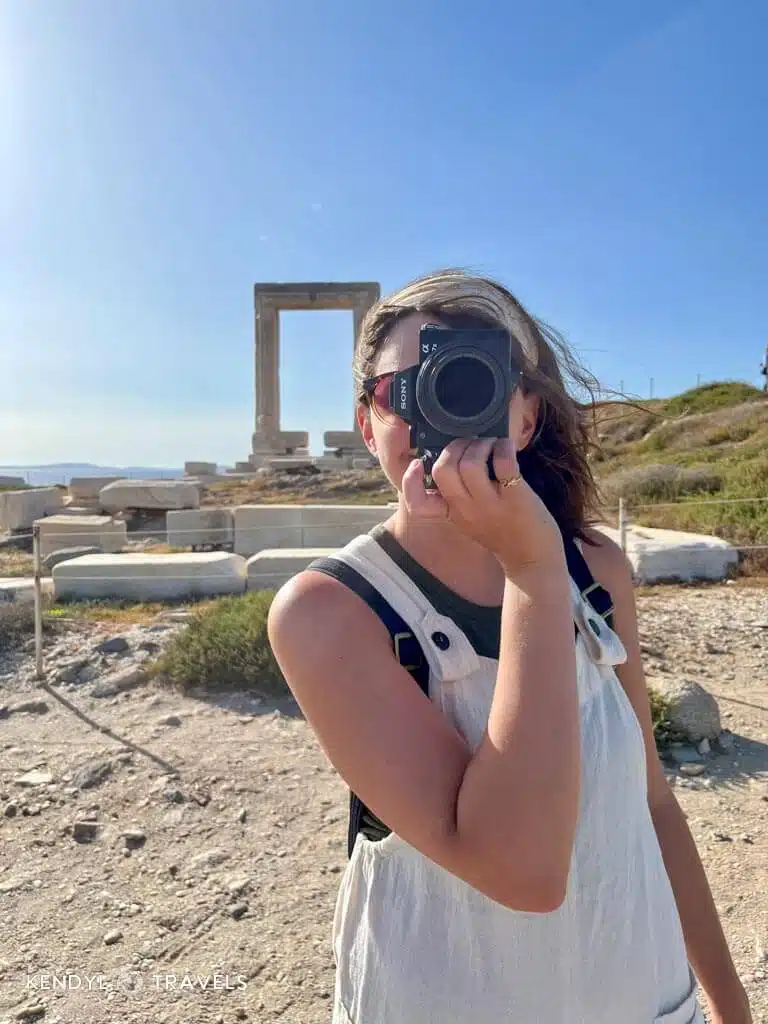 Traveler in white overalls taking photos at the Portara ruins in Naxos, Greece – lightweight layers for a May trip.