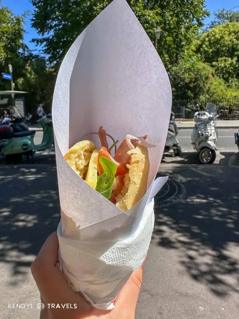 Hand holding a fresh prosciutto and arugula sandwich wrapped in paper on a sunny Paris street.