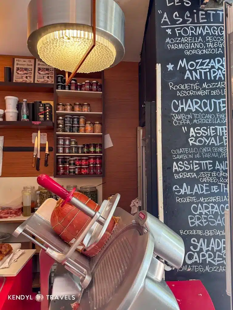 Inside a small Parisian deli with a meat slicer and chalkboard menu listing Italian-inspired dishes.